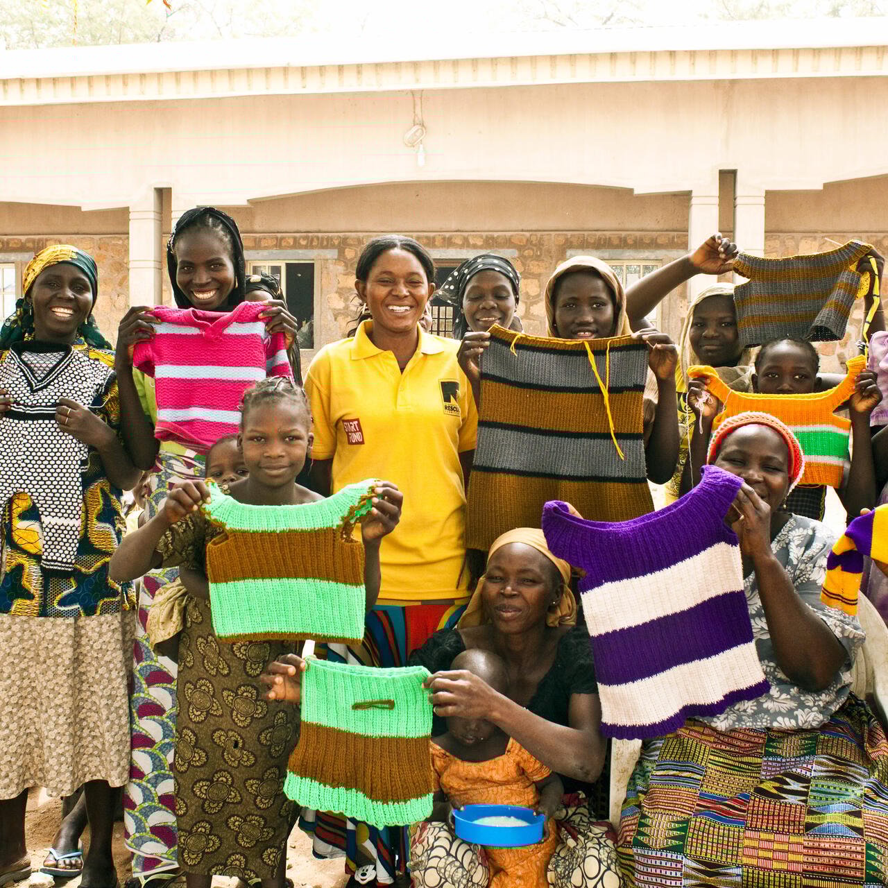 A group of women who are part of an IRC livelihoods program in Nigeria stand outside, lifting hand-knitted tops to show them off.