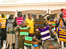 A group of women who are part of an IRC livelihoods program in Nigeria stand outside, lifting hand-knitted tops to show them off.