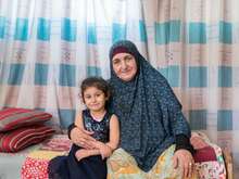A young girl and her grandmother sit at the edge of a bed and pose for a photo.