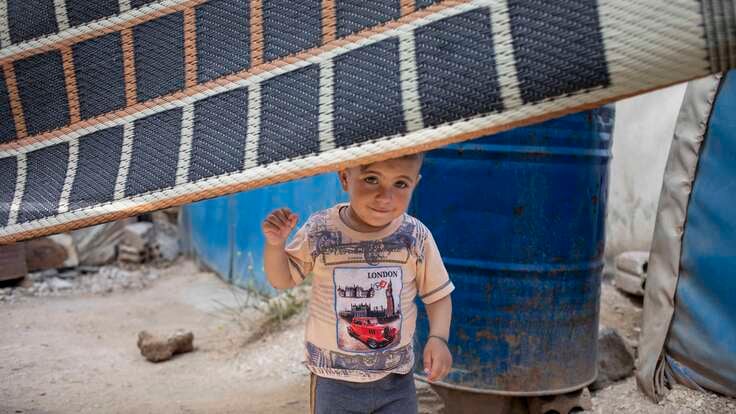 Ein kleines Kind steht in einer provisorischen Unterkunft hinter einer Decke, die als Trennwand dient und hält einen gelben Gegenstand in der Hand.