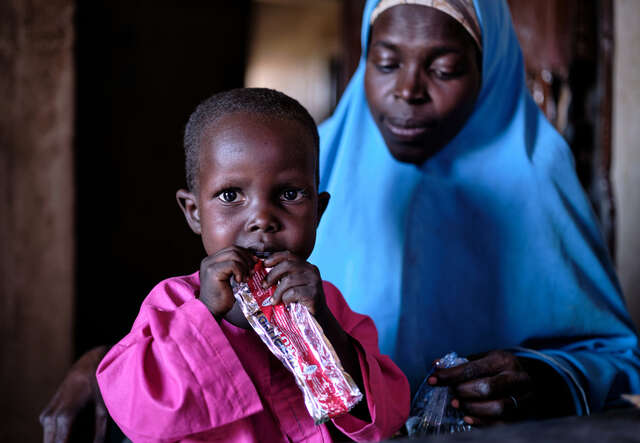 A little boy eating peanut paste as his mother watches him from the background
