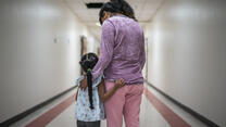 A Mexican mother wearing pink pants and a purple shirt stands with her arms around her young daughter, who has a long braid, in a hallway. They are asylum seekers in an IRC welcome center.