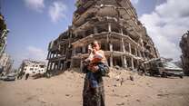 A mother holds a young child in her arms while walking past a destroyed building in Gaza.