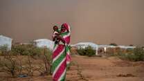 A Sudanese mother holds her young child in her arm. They stand on the outskirts of a camp for displaced people.