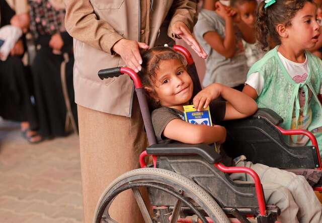 A young Palestinian girl in a wheelchair holds a pack of colored pencils in her arms and smiles.