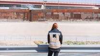 An IRC staff member faces away from the camera and towards the Mexico - U.S. border, while adorning a vest that has the IRC logo on the back.